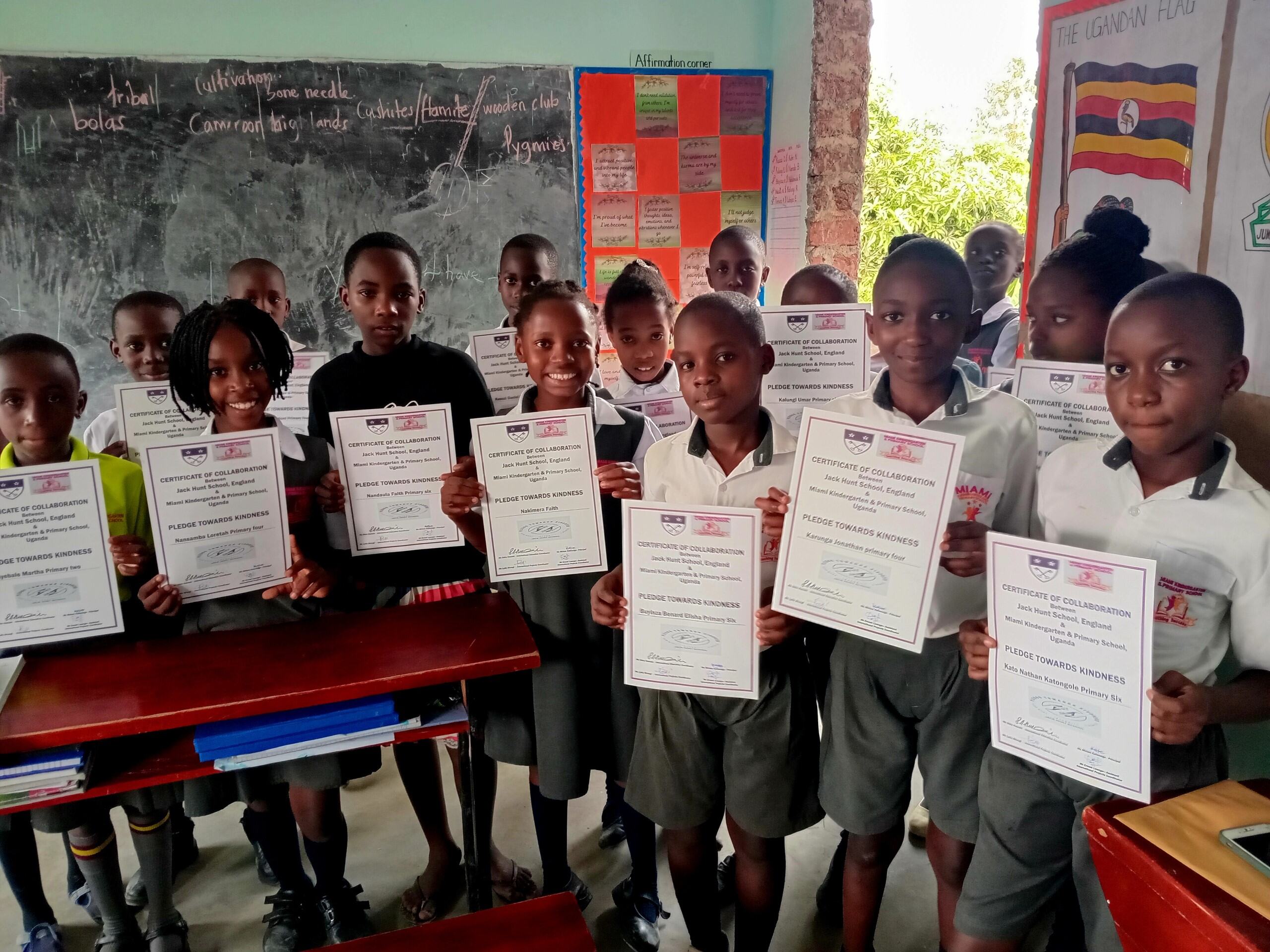 Students from Miami Kindergarten and Primary School in Uganda stood holding certificates of collaboration for Pledge Towards Kindness